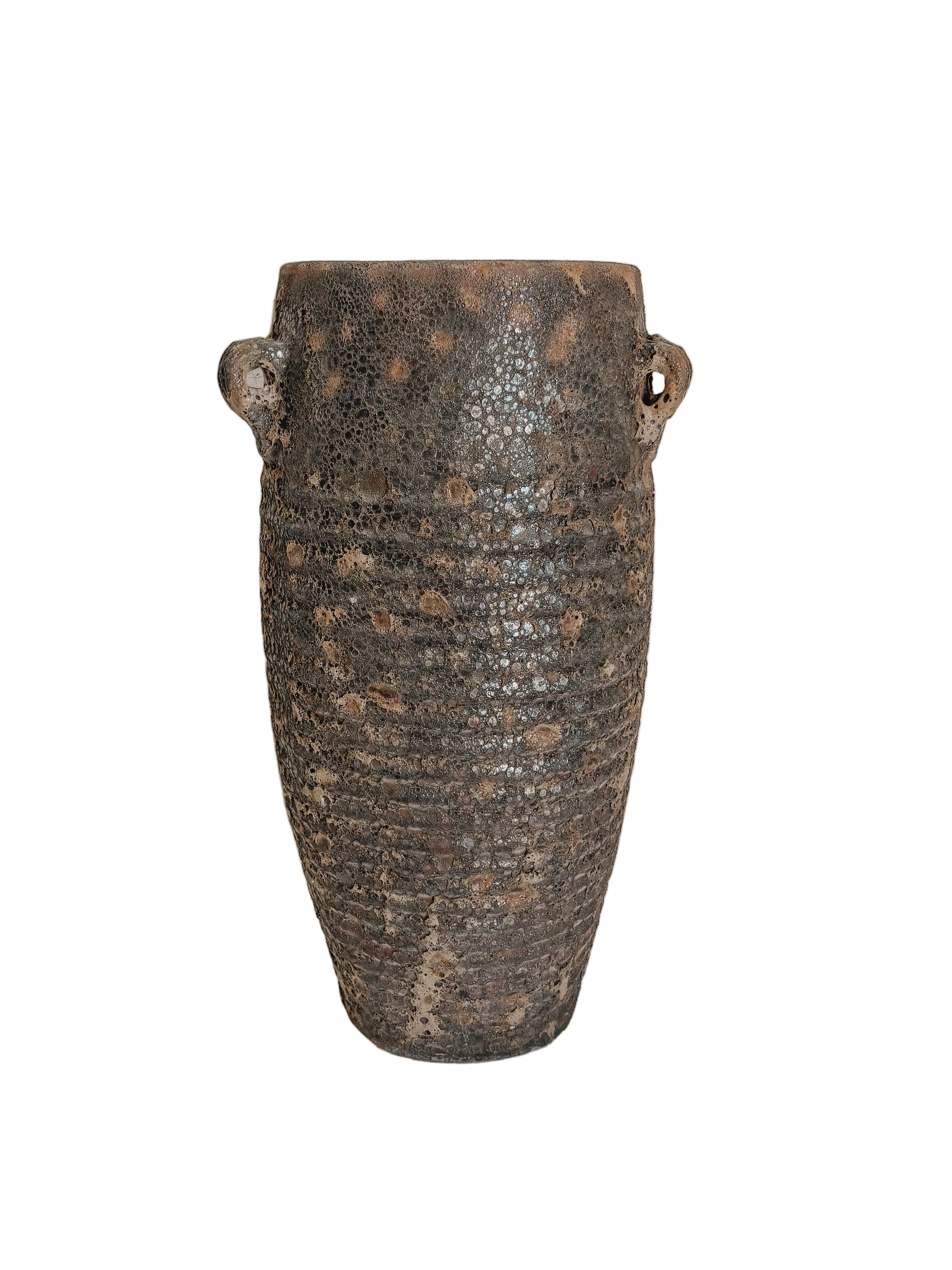 A rustic dark brown ceramic vase with a textured, speckled glaze and two small loop handles, displayed on a white pedestal against a neutral beige wall.