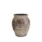 A small rustic ceramic pot with a distressed, mottled glaze in earthy brown and off-white, displayed on a white pedestal against a neutral beige wall.