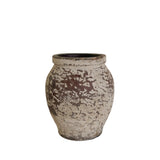 A small rustic ceramic pot with a distressed, mottled glaze in earthy brown and off-white, displayed on a white pedestal against a neutral beige wall.