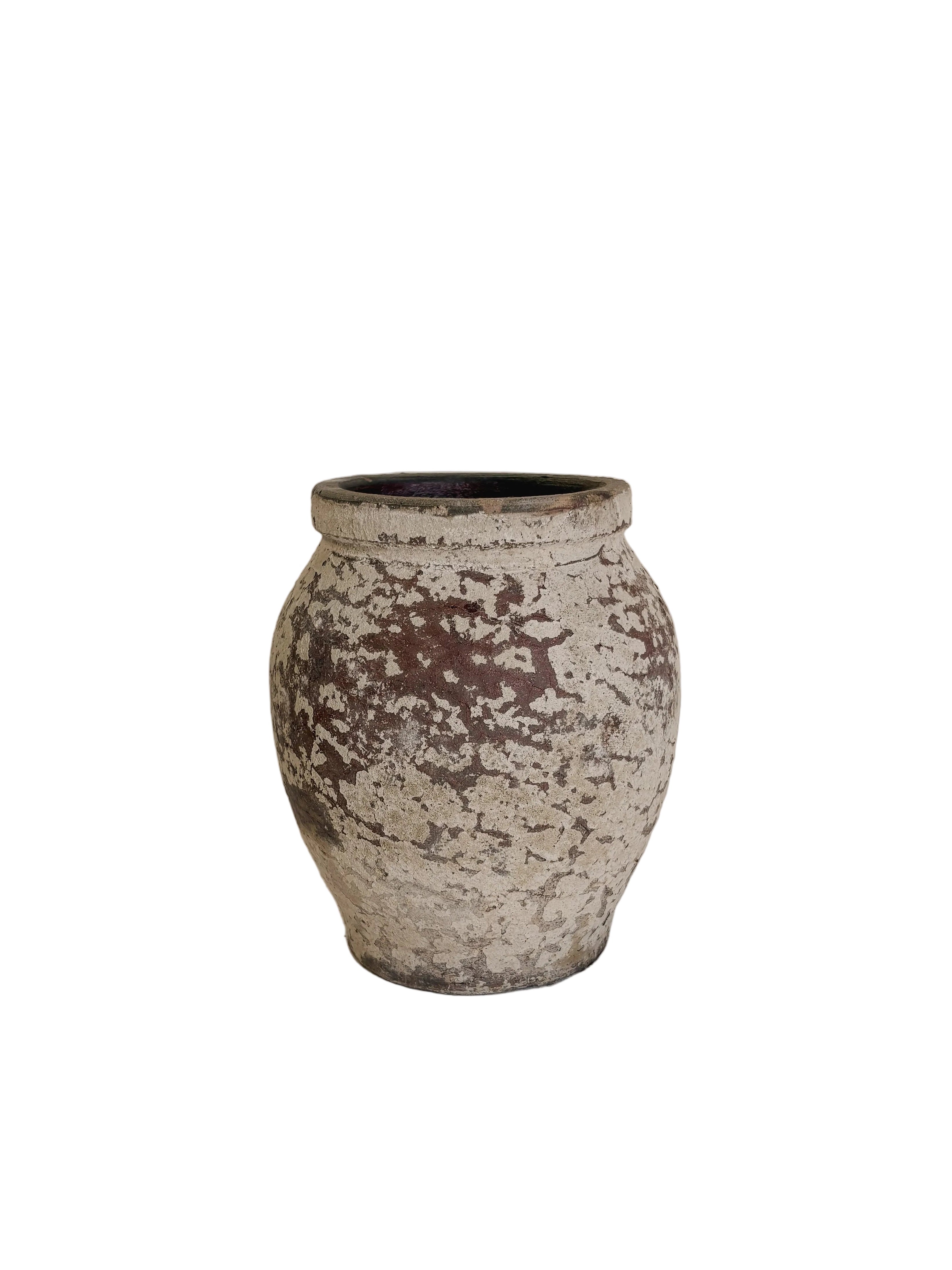 A small rustic ceramic pot with a distressed, mottled glaze in earthy brown and off-white, displayed on a white pedestal against a neutral beige wall.