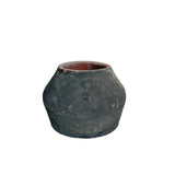 A small rustic dark green ceramic pot with a terracotta-colored interior, displayed on a light gray pedestal against a neutral beige background.